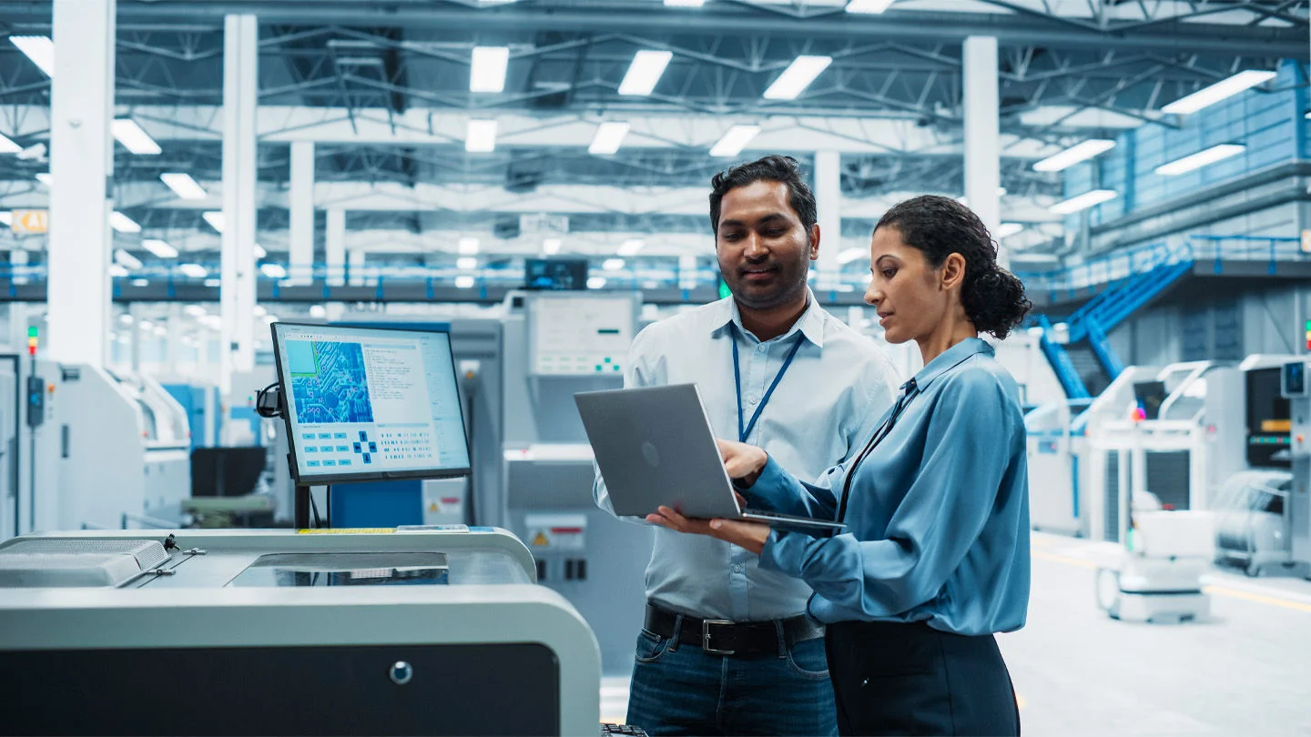 Two professionals stand in a high-tech manufacturing environment, reviewing data on a laptop and a nearby monitor.