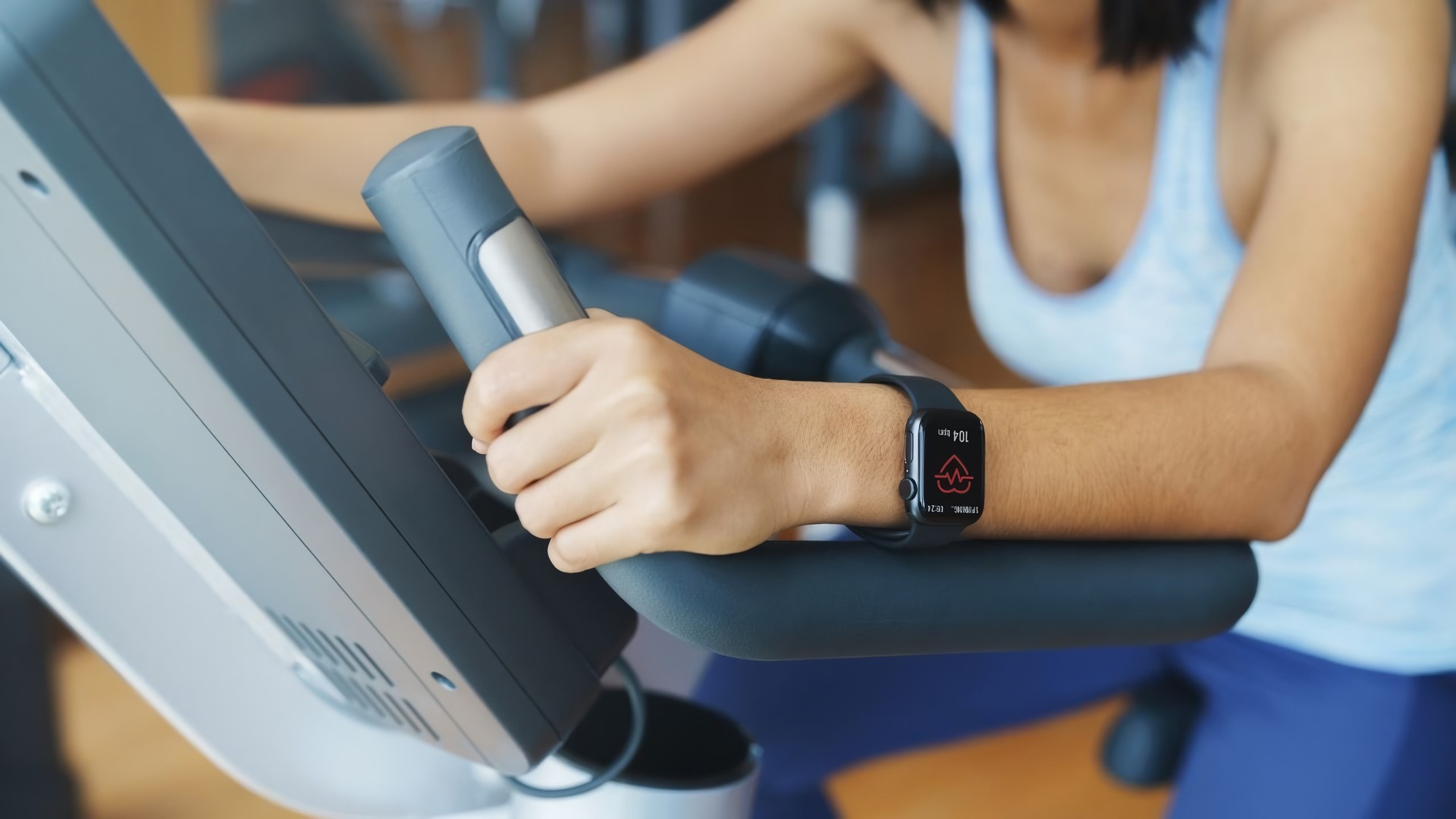 A young woman wearing a smartwatch is engaged in a cycling workout on a stationary bike in a gym setting